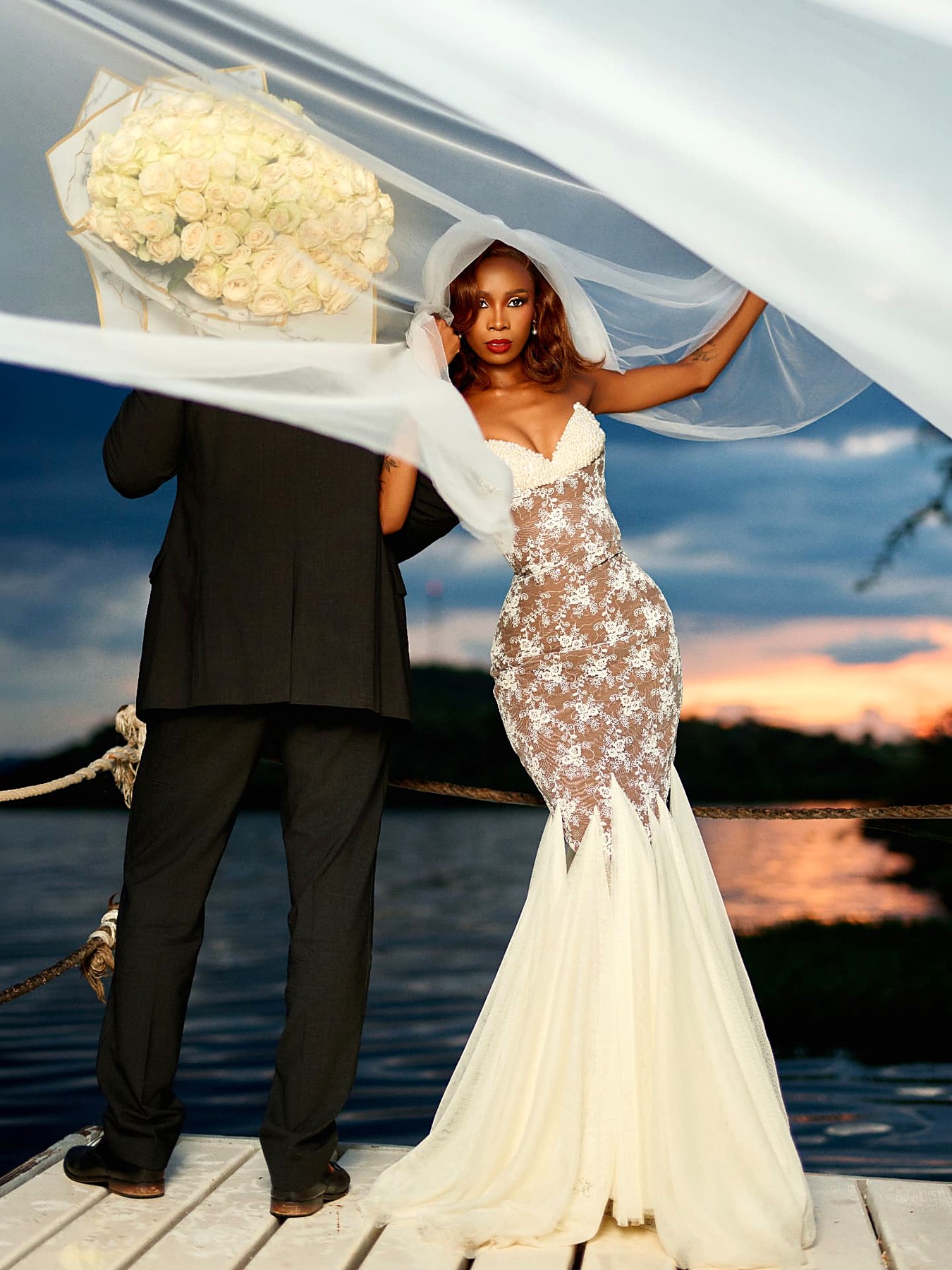 Bride and groom on a jetty at sunset, her lace mermaid gown and veil lifted by the wind